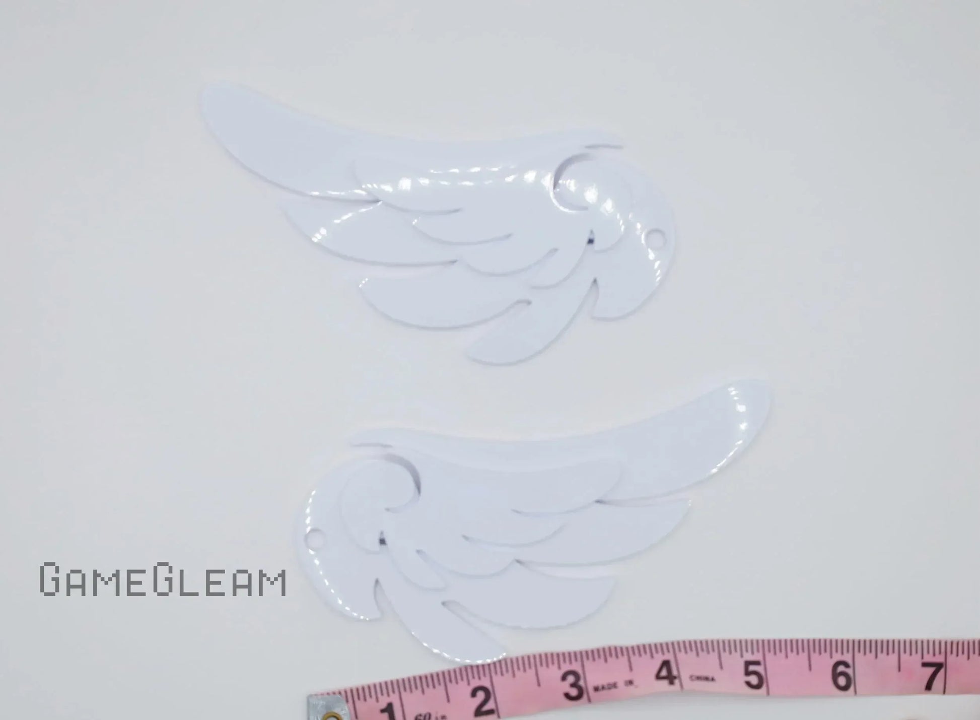 Pair of white acrylic wing-shaped charms with cutouts, shown next to a measuring tape
