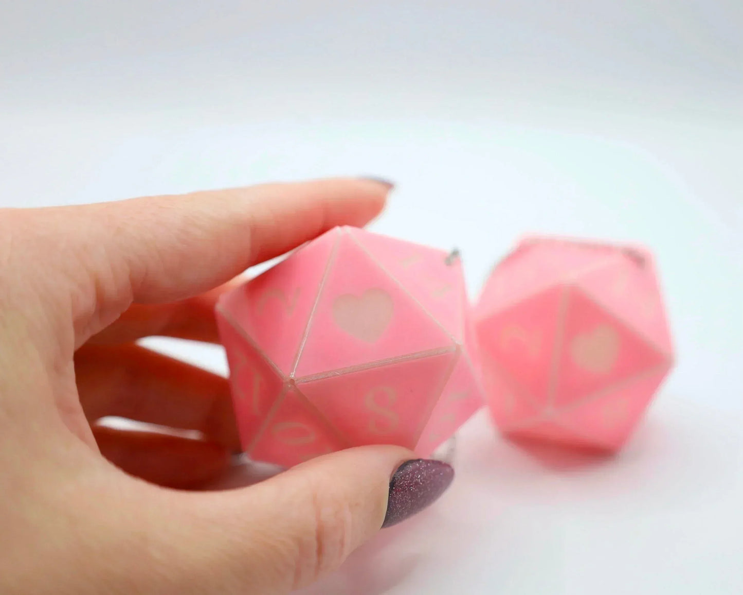 Hand holding a pink 20-sided dice charm with heart and number symbols, geek car accessory