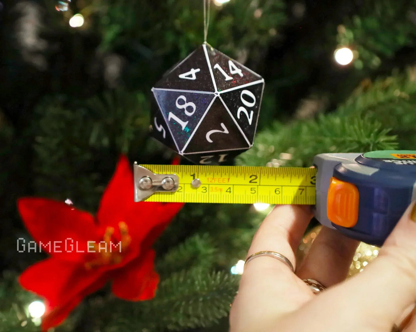 Black 20-sided dice ornament measured with tape, hanging on Christmas tree with red flower.