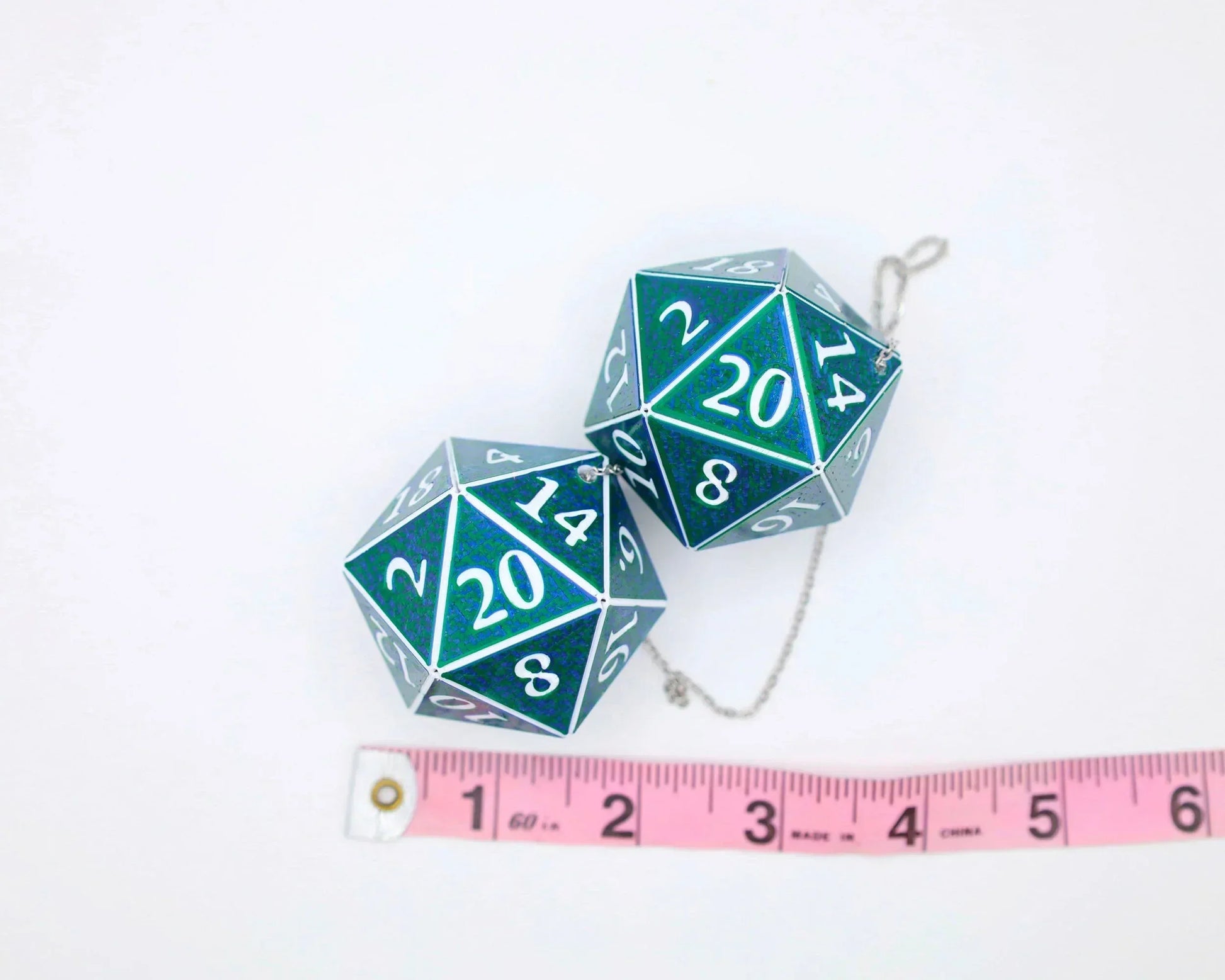 Pair of teal holographic 20-sided dice on a chain, shown with a pink measuring tape