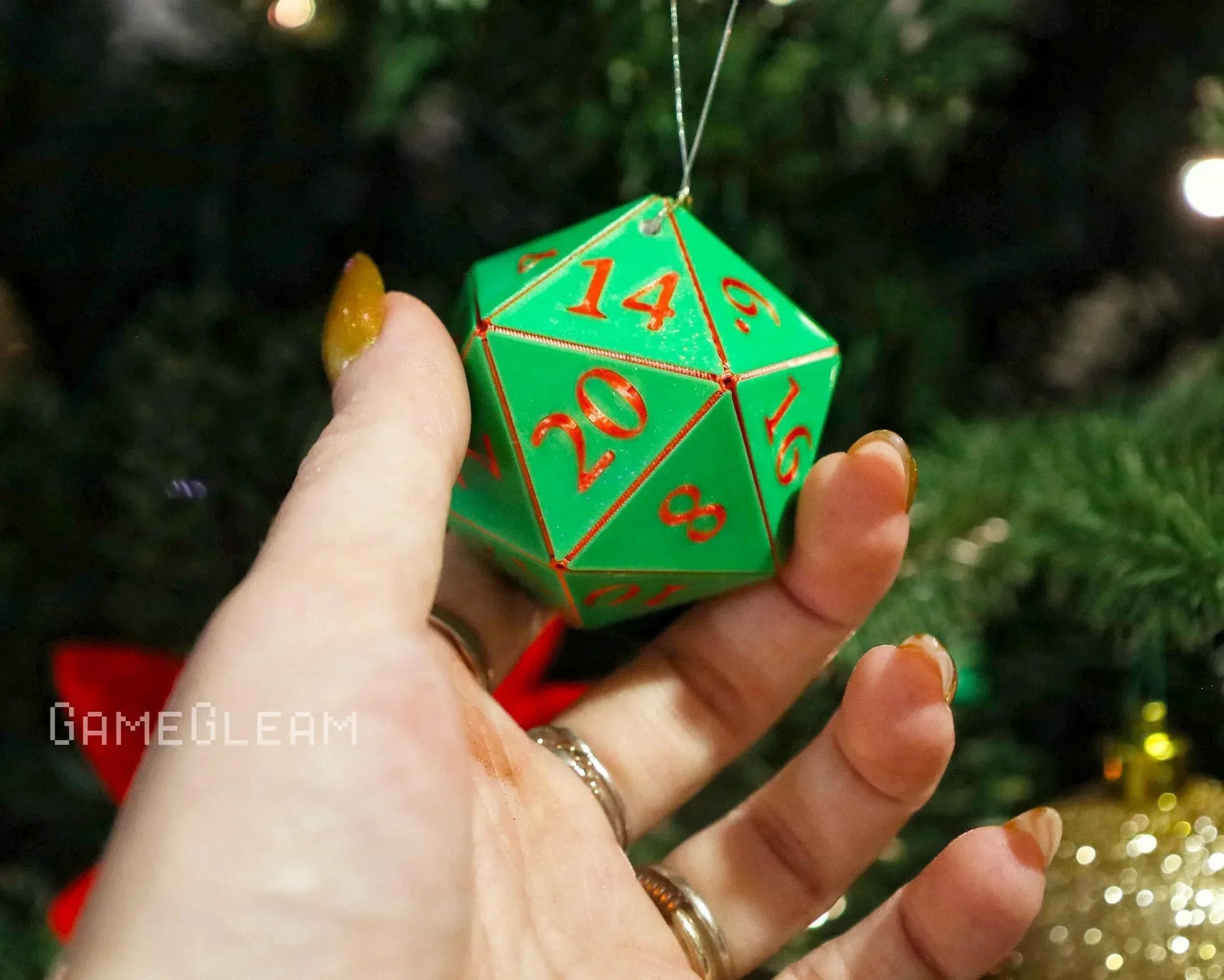 Hand holding a green twenty-sided dice ornament with red numbers near a Christmas tree.
