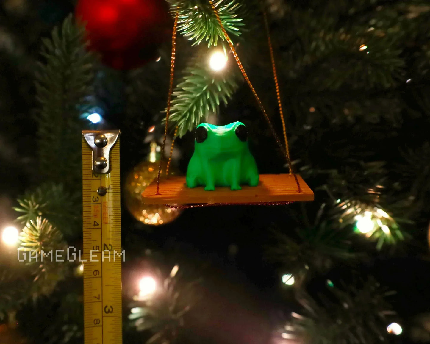 Handmade green frog ornament on swing, hanging on Christmas tree with measuring tape for scale