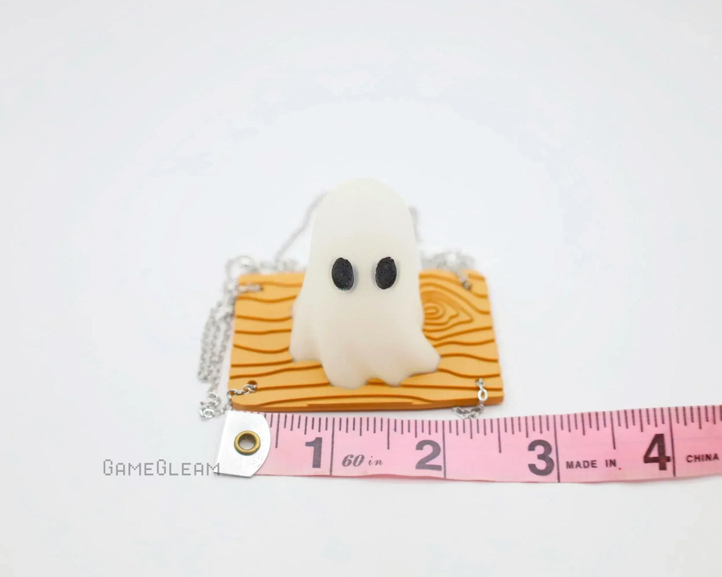 Handmade ghost charm with black eyes on textured orange base, next to a pink measuring tape
