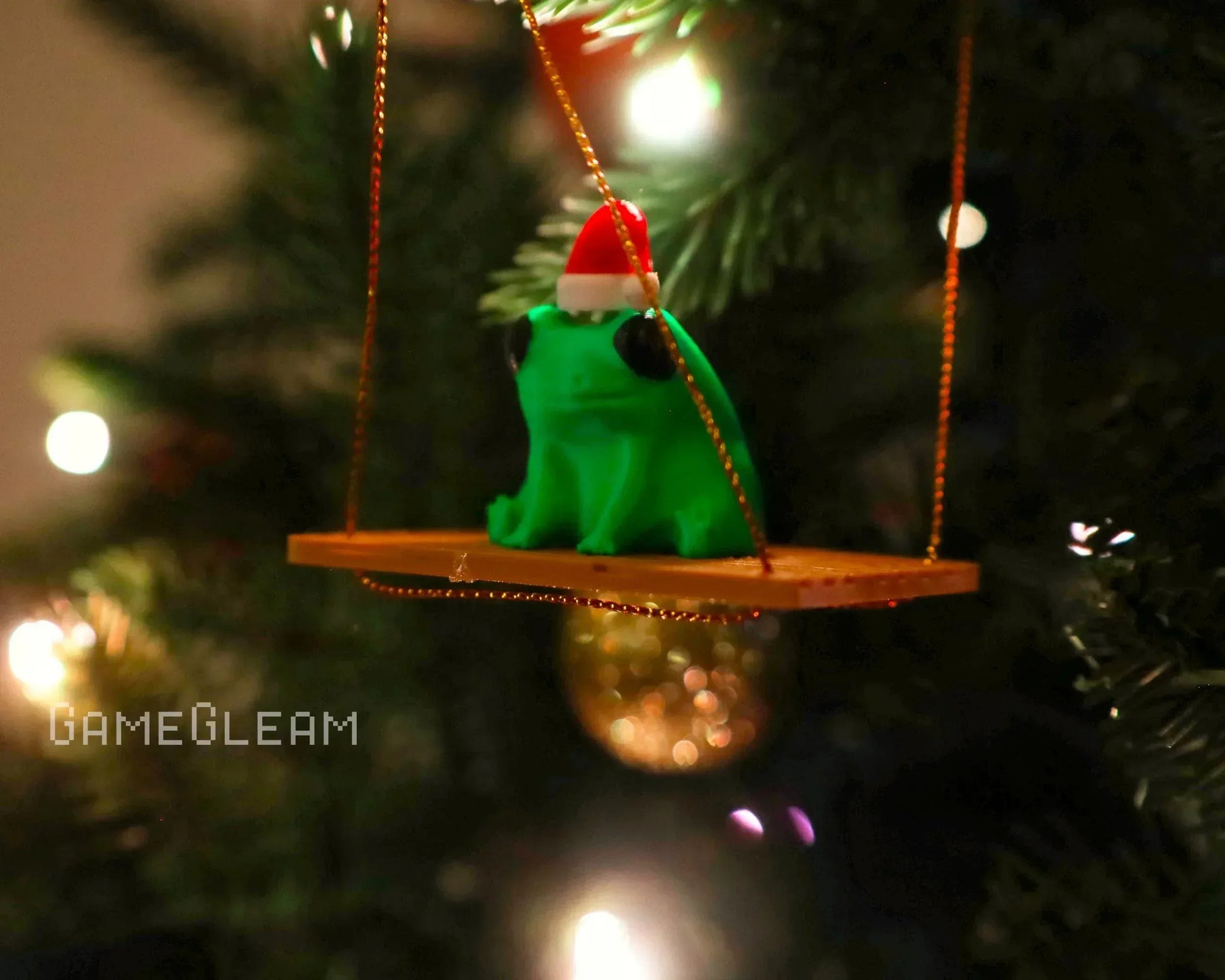 Green frog ornament with Santa hat on swing, hanging on a Christmas tree with lights.