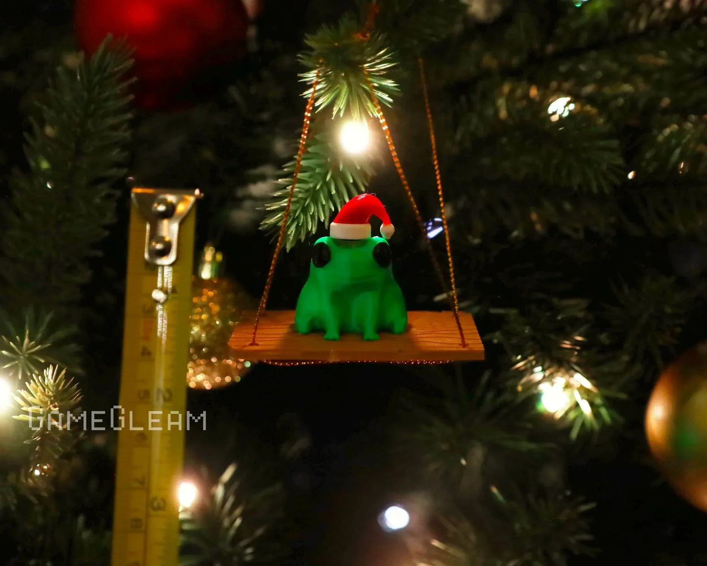 Green frog ornament with Santa hat on a swing, hanging on a lit Christmas tree
