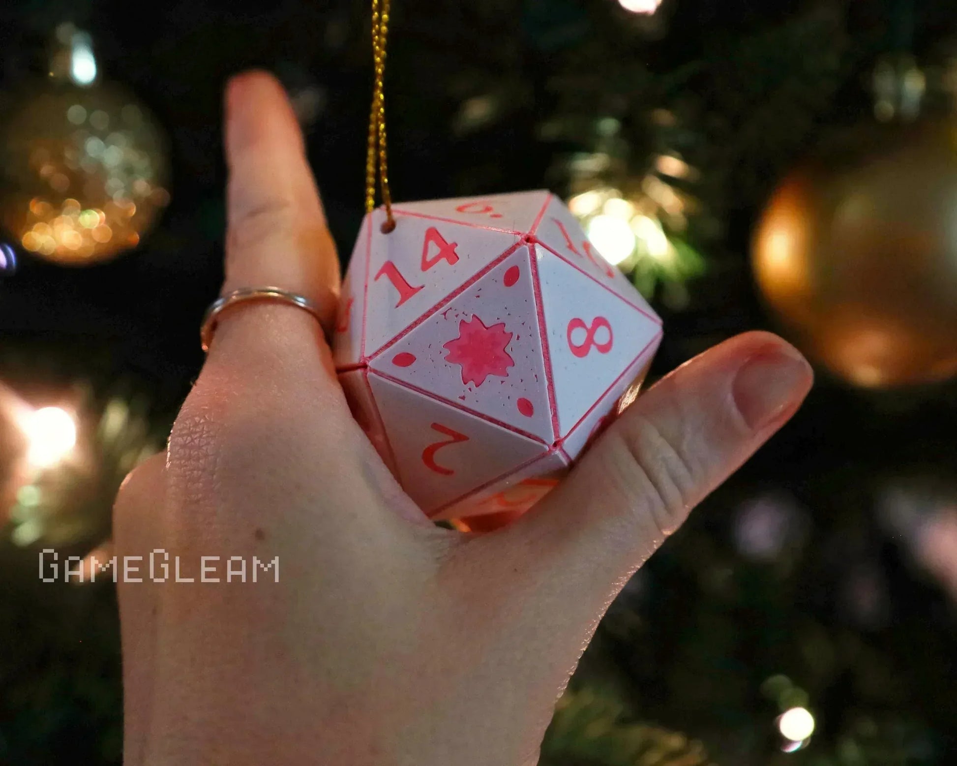 Hand holding hanging pink twenty-sided dice ornament with numbers, near Christmas tree