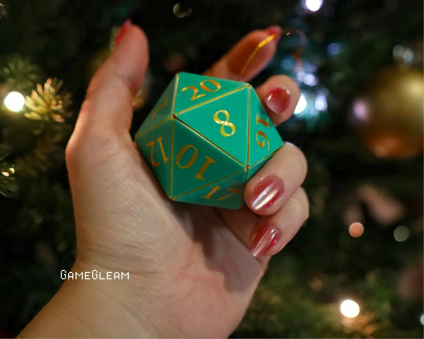 Hand holding teal twenty-sided dice ornament with gold numbers near Christmas tree