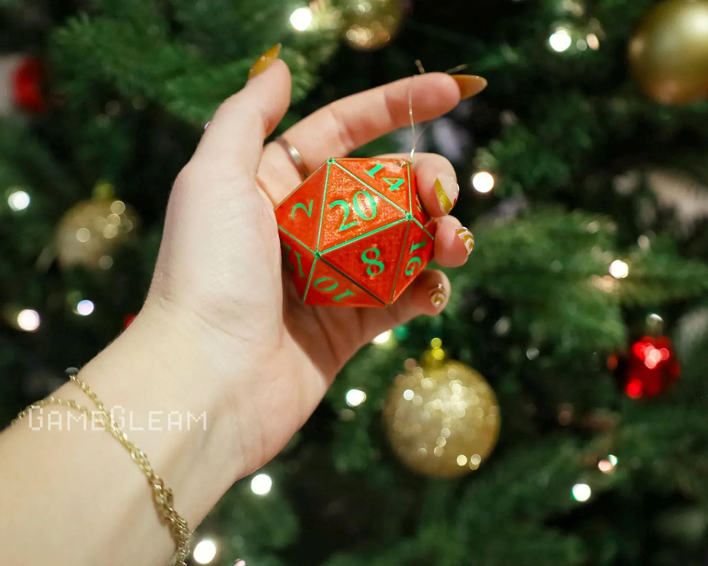 Hand holding a red twenty-sided polyhedral dice ornament in front of a decorated Christmas tree.